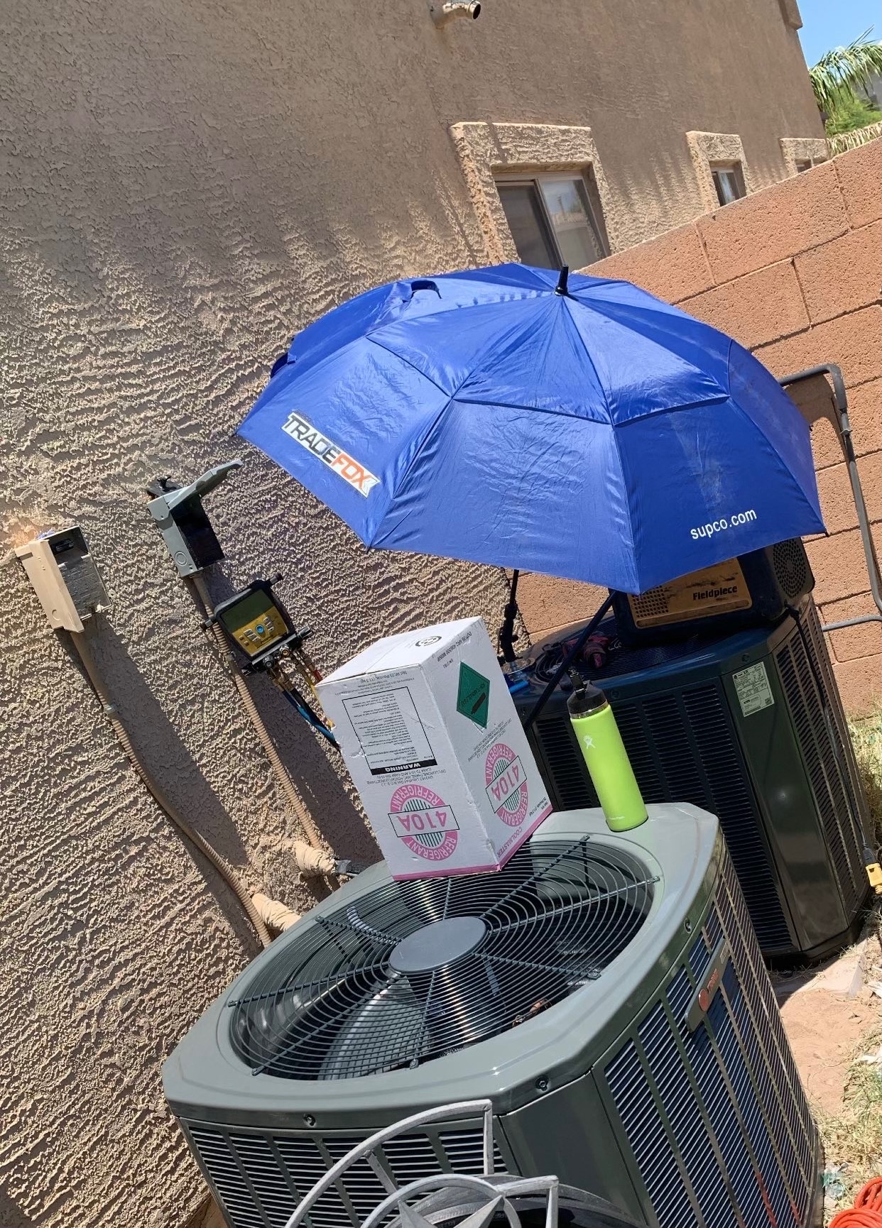 Two air conditioning units outside a residential home undergoing HVAC maintenance. A blue technician's sunshade is set up for shade, with a refrigerant box and