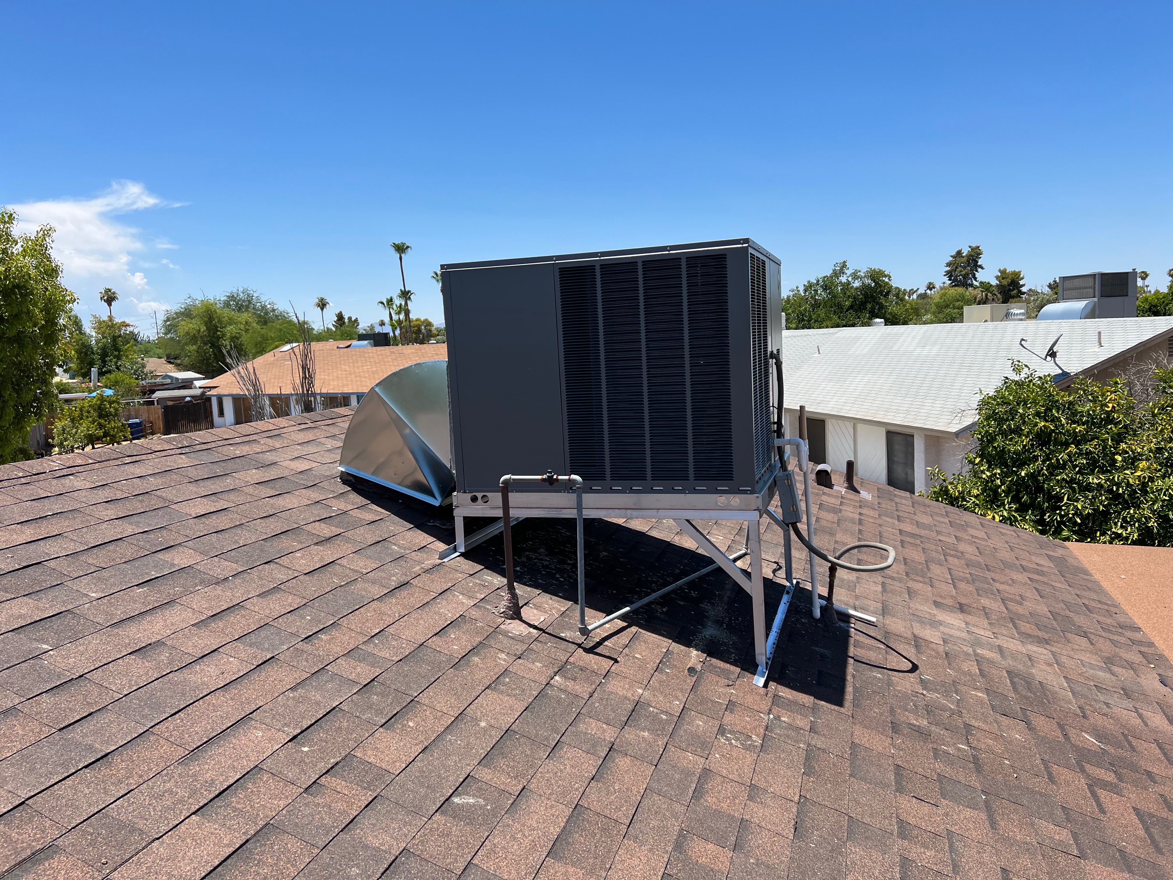 A rooftop HVAC package unit installed on a residential home, elevated on a metal stand for stability and efficiency. The surrounding neighborhood and clear blue